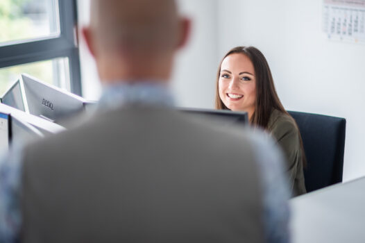 Mitarbeiterin sitzt am Schreibtisch und schaut lächelnd zum Gesprächspartner.