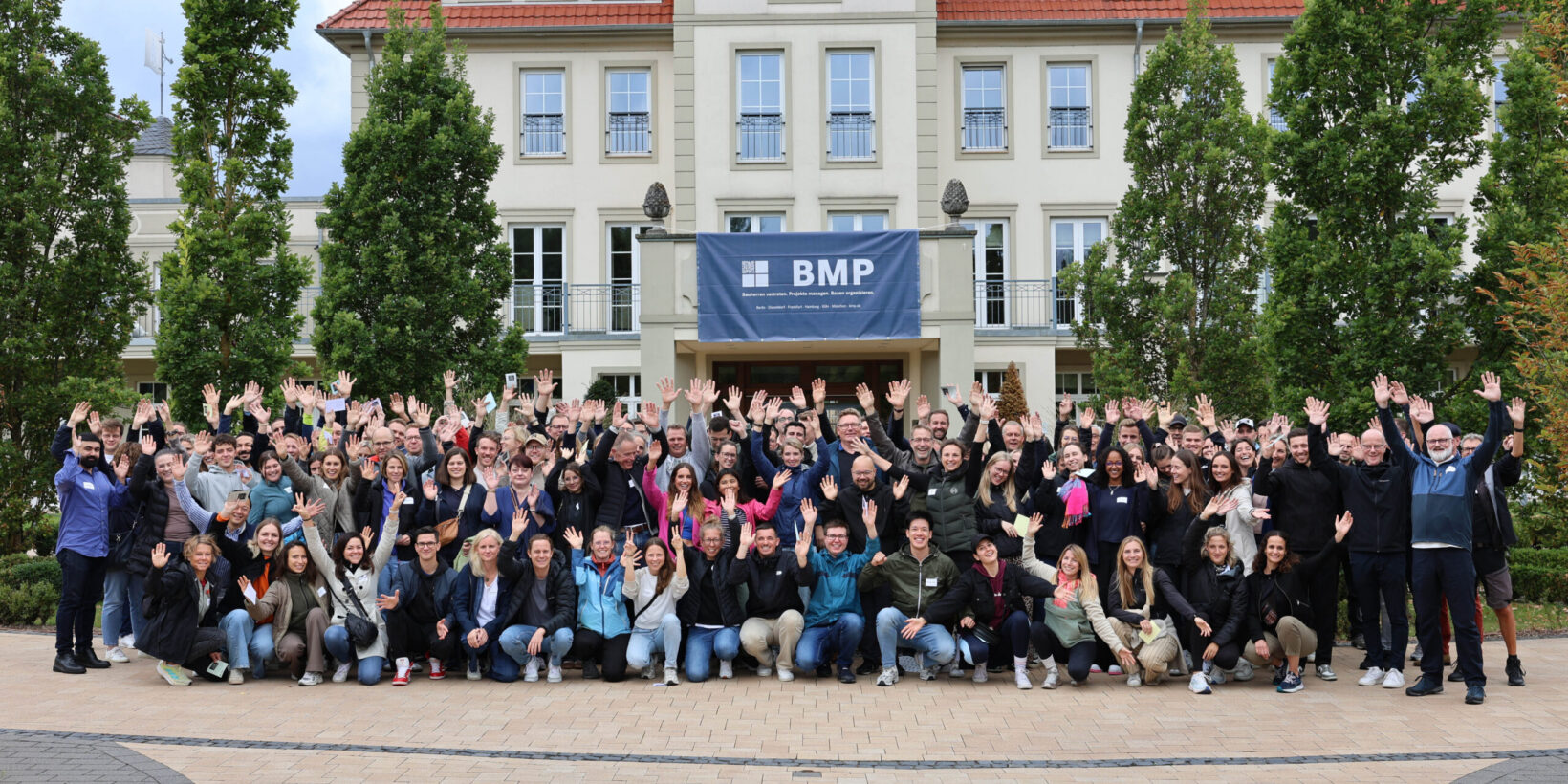 BMP-Team winkt beim Gruppenfoto vor Gebäude mit BMP-Banner.