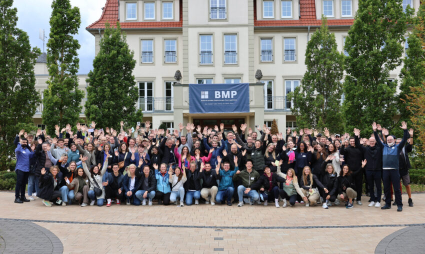 BMP-Team winkt beim Gruppenfoto vor Gebäude mit BMP-Banner.