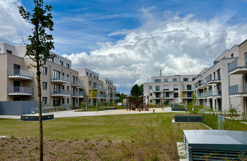 Moderne Wohnanlage mit begrüntem Innenhof, Spielplatz und Sitzbänken unter blauem Himmel.