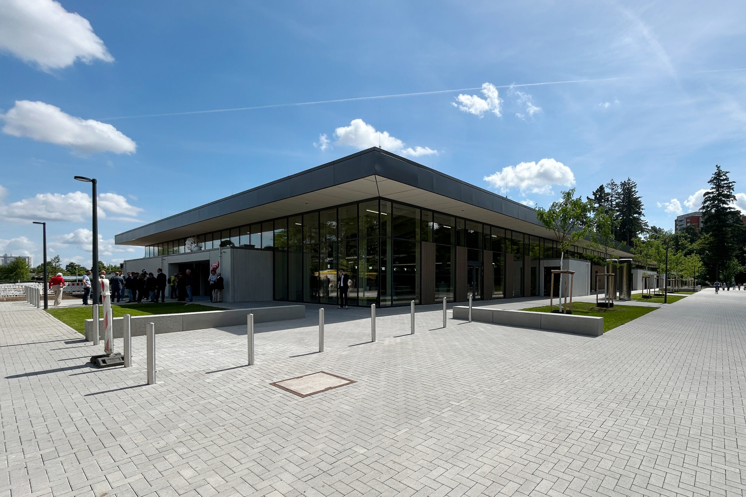 Moderne, eingeschossige Sporthalle mit großer Glasfassade und auskragendem Dach im Schulzentrum Südwest in Karlsruhe-Oberreut. Auf dem Vorplatz mit Pflasterflächen, Pollern und jungen Bäumen stehen Besucherinnen und Besucher während der Einweihung.