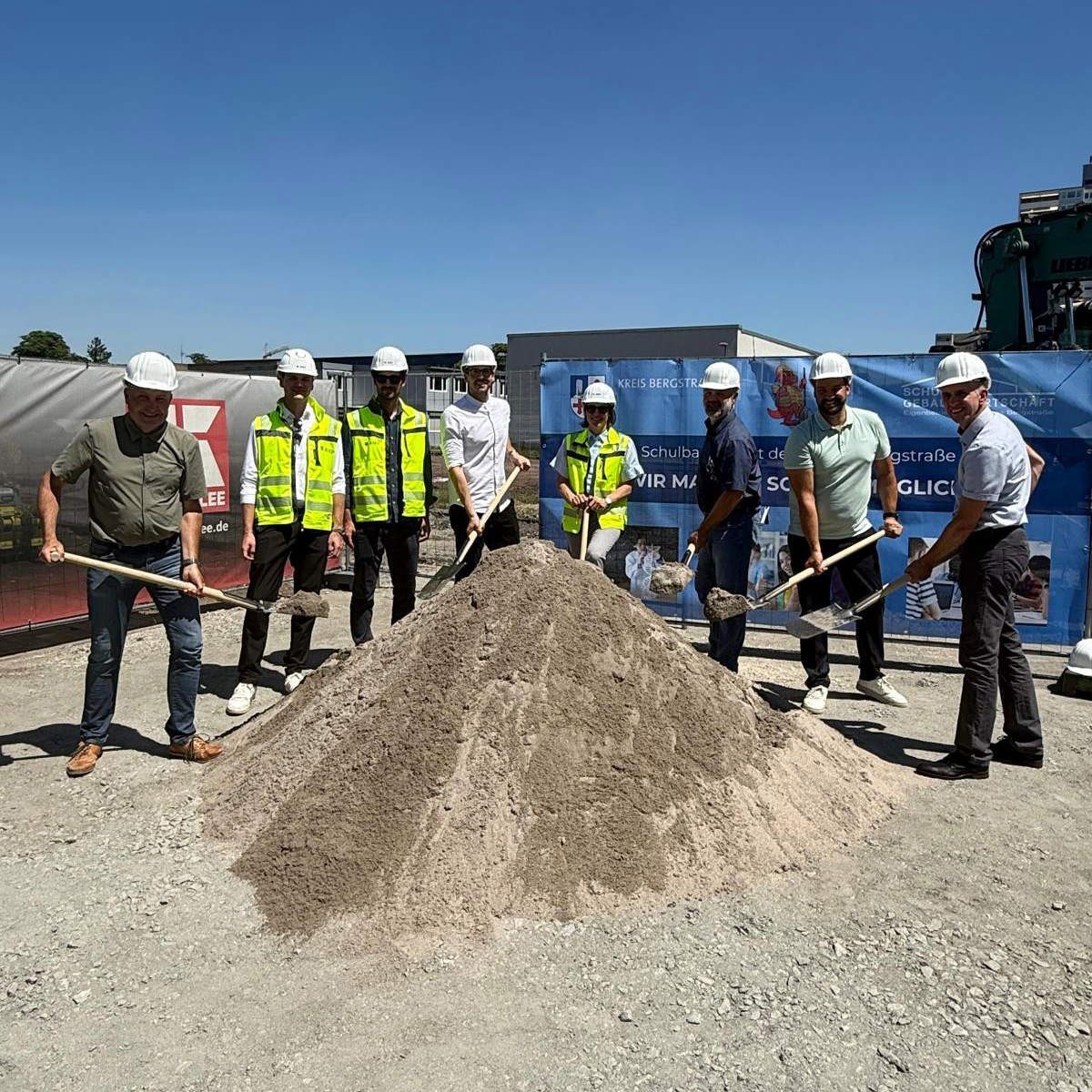 Projektbeteiligte vor einem Sandberg mit einer Schaufel in der Hand beim Spatenstich für das Lessing-Gymnasium.