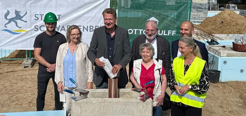Mehrere Personen stehen bei einer Grundsteinlegung im Freien vor einem Bauzaun; eine Person hält eine Maurerkelle, im Hintergrund sind Banner und Gebäude zu sehen.