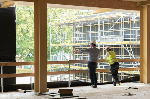 Zwei Mitarbeitende mit Schutzhelmen besichtigen eine Baustelle im Rohbau; im Hintergrund sind ein mehrstöckiges Gebäude mit Gerüst und eine begrünte Umgebung zu sehen.