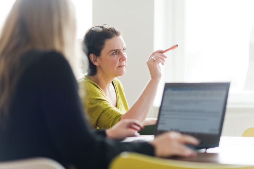 Zwei Mitarbeitende von BMP besprechen gemeinsam ein Projekt; eine Person zeigt mit einem Stift auf etwas außerhalb des Bildausschnitts, während die andere am Laptop arbeitet.